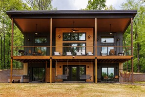 Une maison moderne à deux étages, dotée d’une terrasse en bois, d’un espace salon extérieur et de grandes fenêtres.