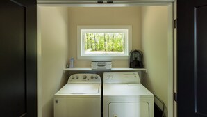 Une buanderie équipée de deux machines à laver et de deux sèche-linge à chargement frontal, d’une fenêtre donnant sur des arbres et d’une étagère sur laquelle se trouvent une radio et une bouteille.