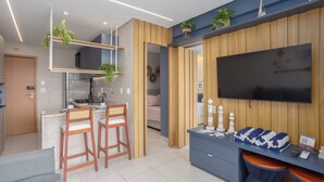 A modern kitchen with a built-in stove, two wooden bar stools, and a large flat-screen TV mounted on the wall. - Mare Nostrum - Carneiros Beach - Ocean View - Balcony - 2 Suites - Eco Resort Condominium (Tamandare)