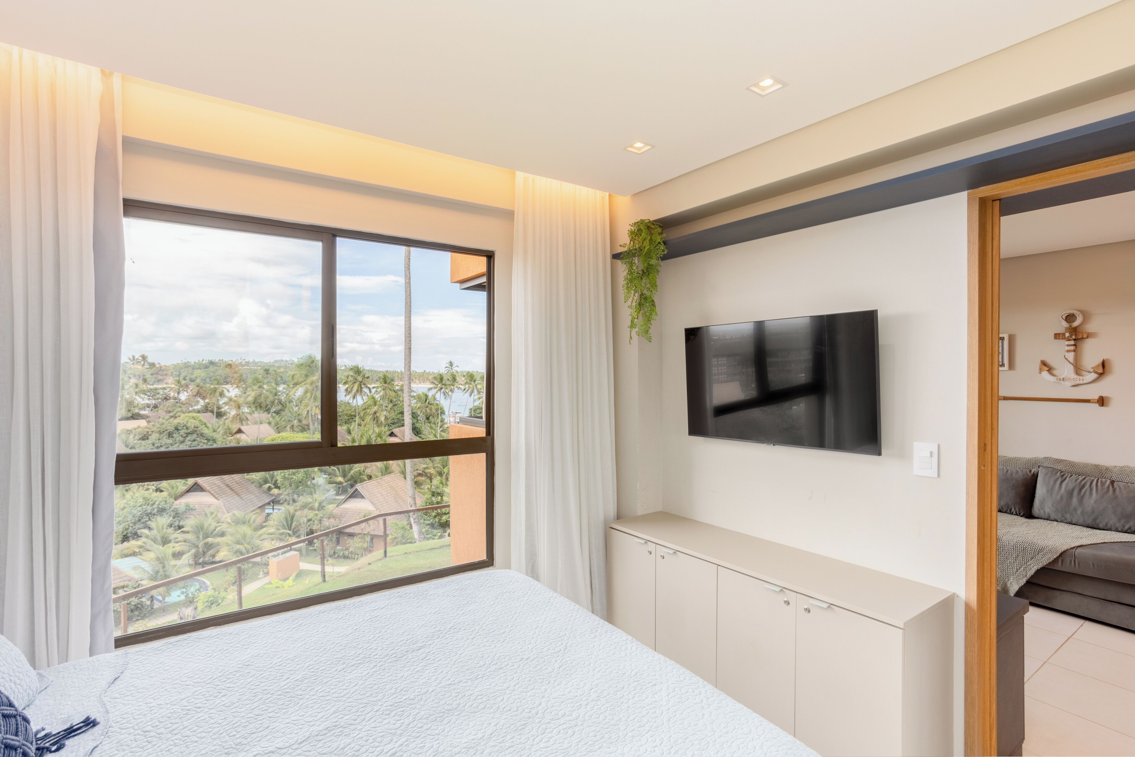 A bedroom with a large window, a bed, a TV mounted on the wall, and a view of the outdoors.