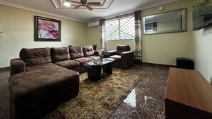 Ein Wohnzimmer mit braunem Ledersofa, einem Glastisch und einem Holznachttisch. Es gibt einen Deckenventilator, ein Fenster mit gemusterten Vorhängen und ein gerahmtes Bild an der Wand.