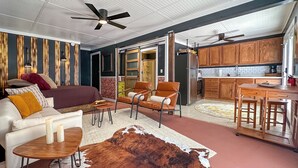 Una sala de estar moderna con un sofá, sillas, un tapete y una zona de cocina con armarios de madera y un refrigerador.