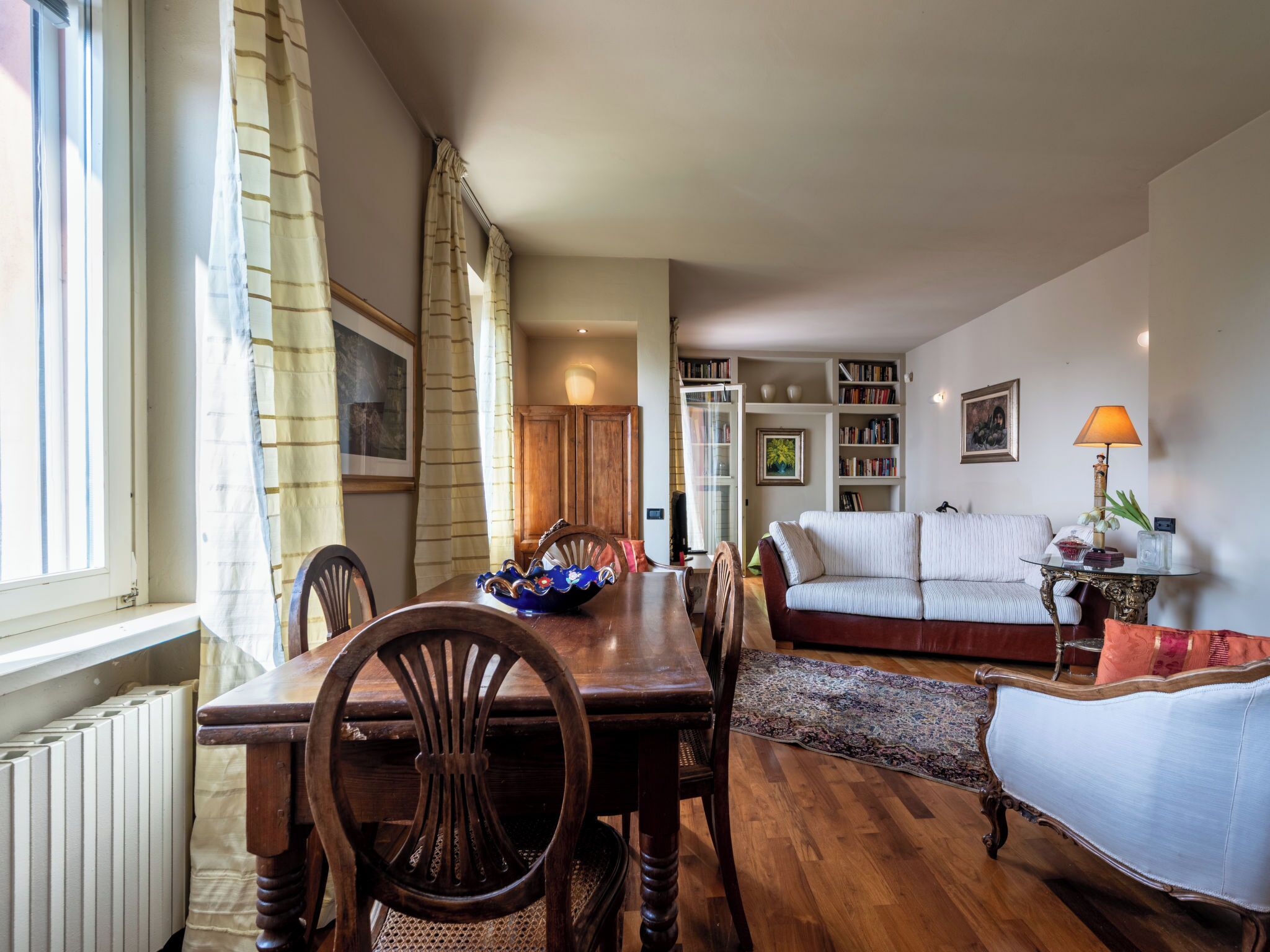 A cozy living room with a dining area, featuring a wooden table, chairs, a sofa, and a bookshelf.