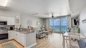 A modern kitchen with a central island, a dining area with a table and chairs, a living area with a sofa, and a large window with a view of the ocean.