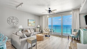 A spacious living room with a large window offering a view of the ocean, a ceiling fan, and a dining area with chairs.