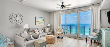 A spacious living room with a large window offering a view of the ocean, a ceiling fan, and a dining area with chairs.