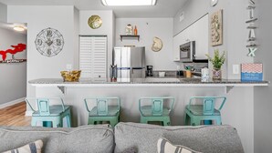 A compact kitchen with a central island, a refrigerator, and a microwave. There are two teal chairs and a sofa with a striped cushion.