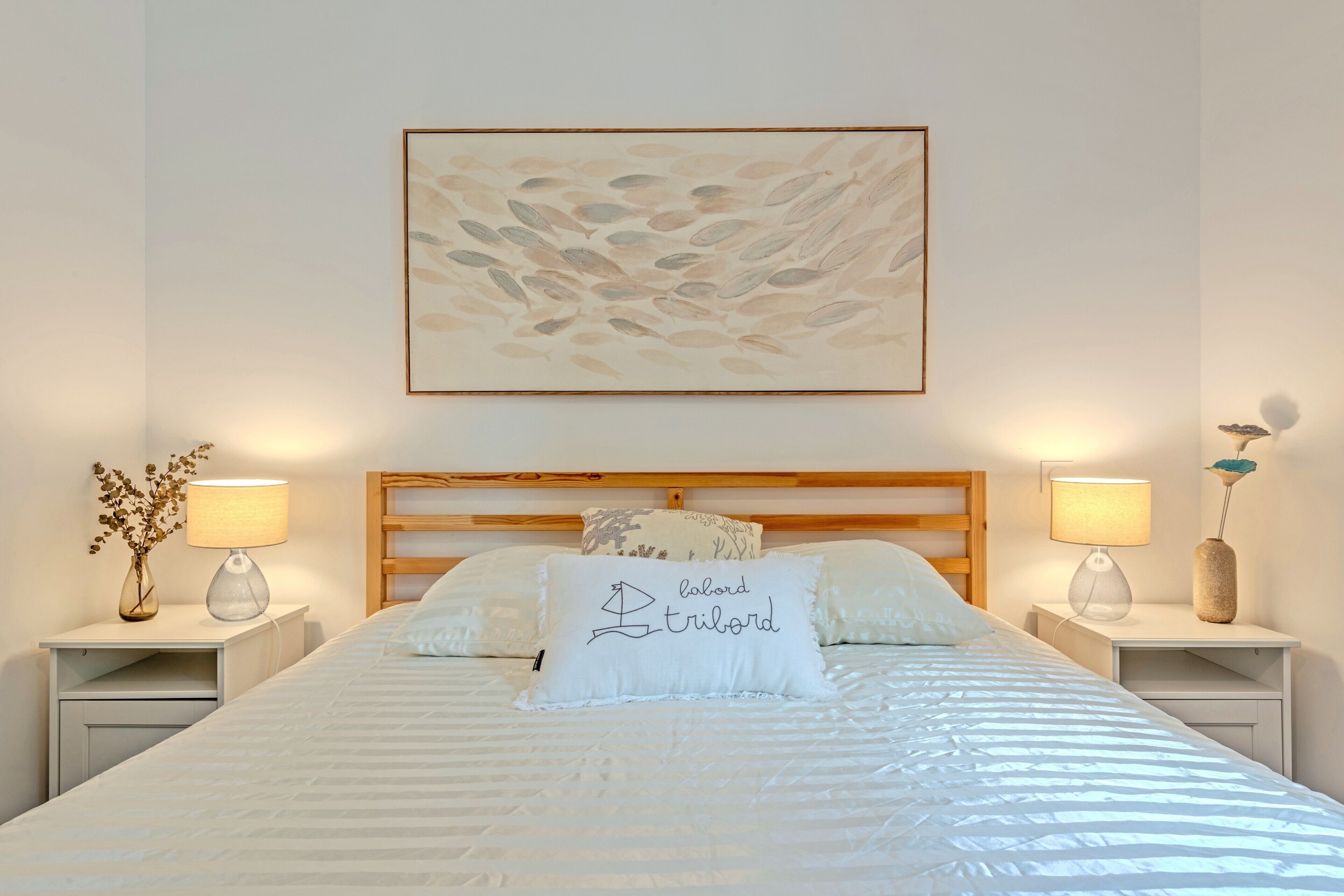 Una cama bien hecha con un cojín que tiene el texto 'lobord tribord', dos lámparas de noche, un cuadro abstracto colgado en la pared y un cabecero de madera.