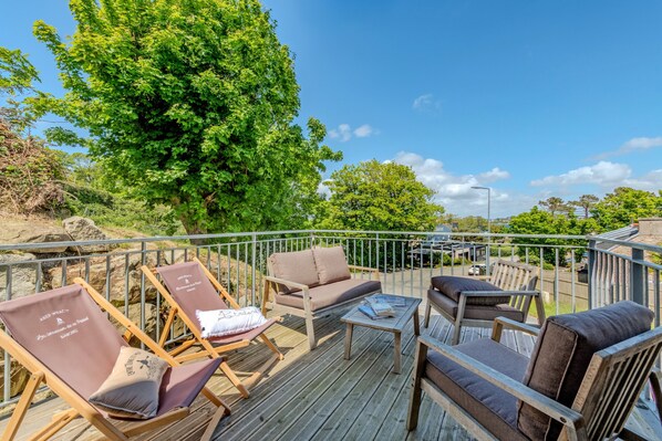 A deck with outdoor seating, including lounge chairs and a sofa, surrounded by greenery and trees.