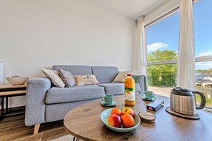 A modern living room with a grey sofa, a wooden coffee table with fruits, a kettle, and a bottle of orange juice.