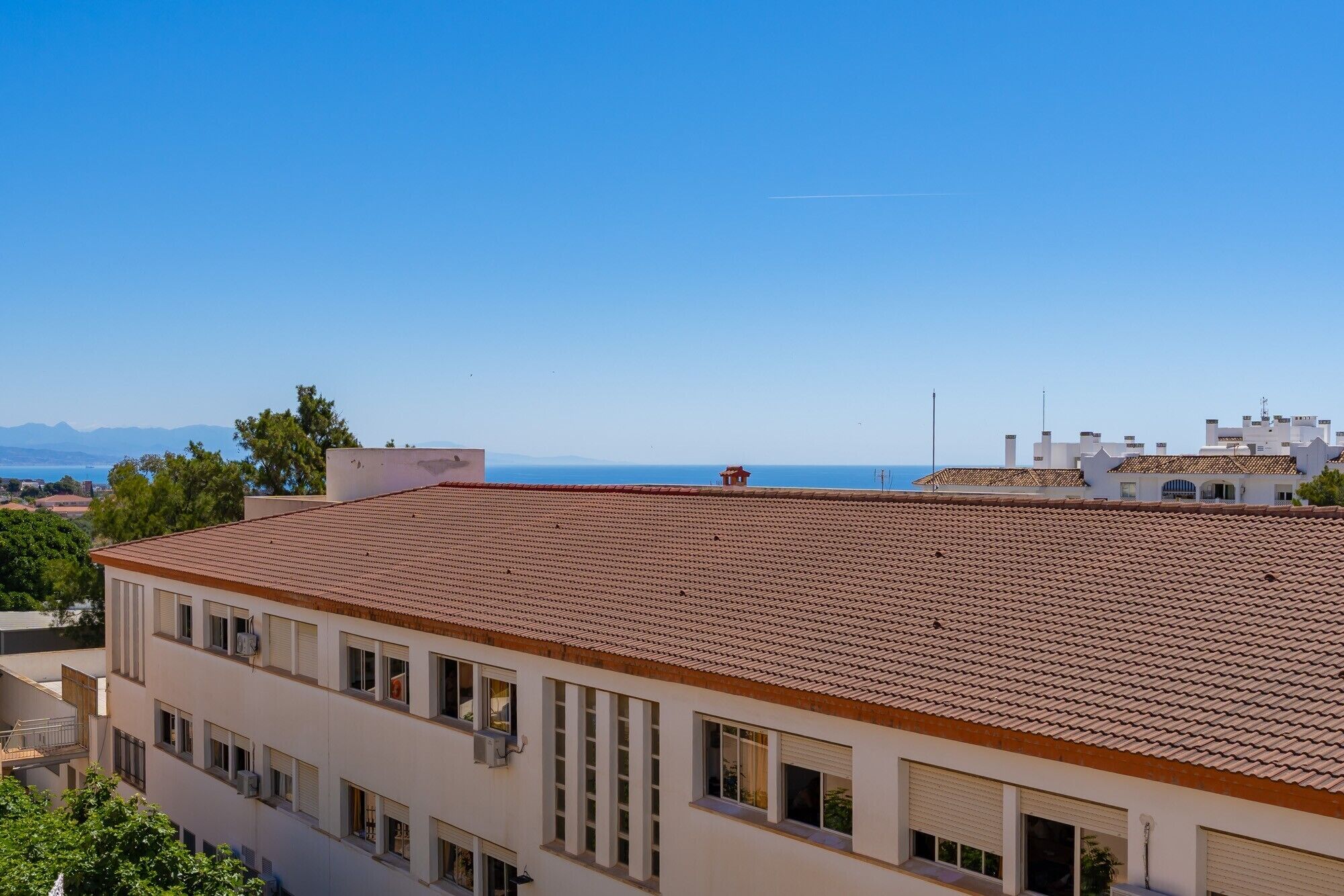 Un edificio con tejado de tejas, varias ventanas y vista al mar y montañas al fondo.