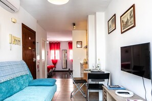 Une pièce comprenant un canapé bleu, un téléviseur, une table à manger avec des chaises et un coin cuisine.