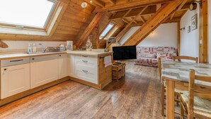 A cozy attic bedroom with a wooden ceiling, a large skylight, a bed with a patterned blanket, a wooden dining table, and a kitchen area with cabinets and a sink. - Apartment 'Passoud 203' with Mountain View, Shared Garden and Wi-Fi (VAUJANY)