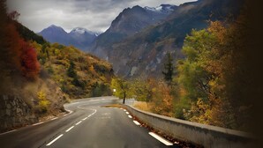 Eine kurvenreiche Straße durch ein gebirgiges Gebiet mit Herbstlaub und in der Ferne schneebedeckten Gipfeln.