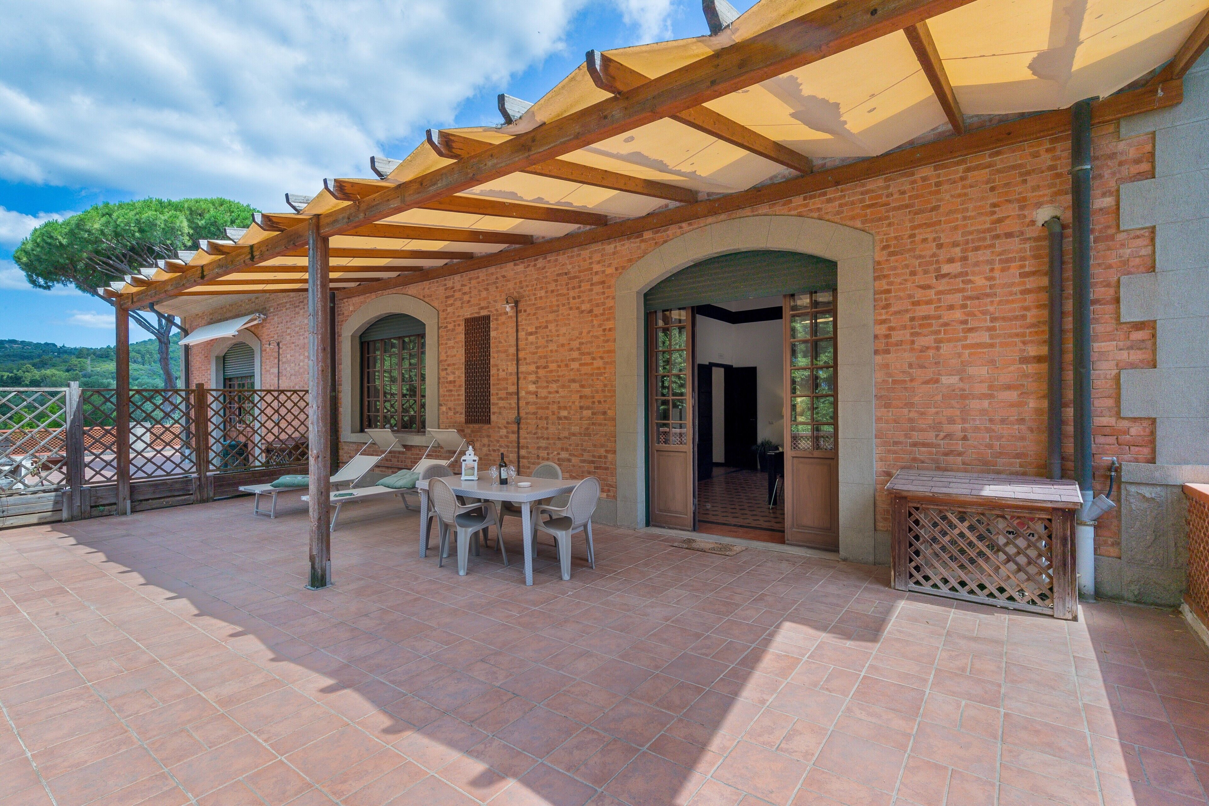 Een overdekt terras met een bakstenen muur, een tafel met stoelen en een houten pergola.