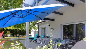 Un patio exterior con un paraguas azul, sillas blancas, una mesa y una parrilla.