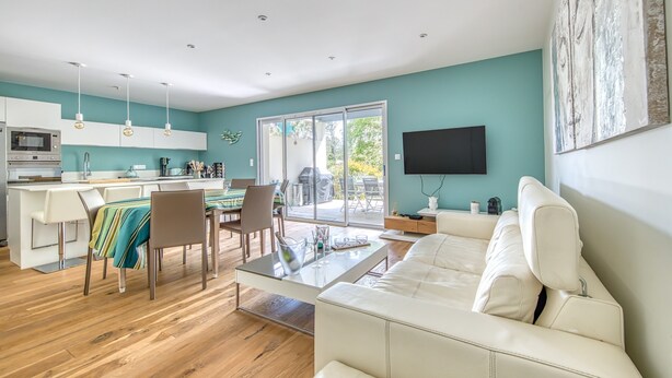 Una cocina moderna con armarios blancos, una zona de comedor con sillas y una mesa, un sofá blanco y un televisor montado en la pared.