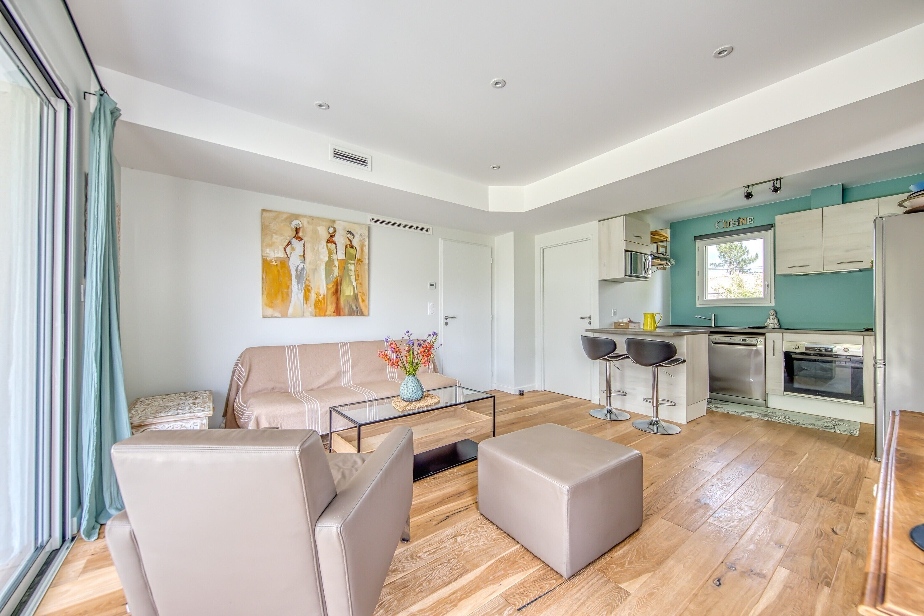 A modern living room with a sofa, ottoman, and coffee table, a kitchen with a dining area, and a large window.