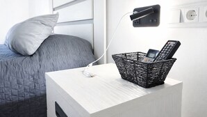 A neatly made bed with a grey quilt, a white pillow, a bedside table with a black woven basket containing a remote control, and a wall-mounted lamp. - Apartment 'Bungalow planta Alta 29' with Shared Pool, Wi-Fi and Air Conditioning (Torrevieja)