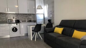 A compact kitchen with a washing machine, a dining area with a chair, and a sofa with yellow cushions. - Apartment 'Bungalow planta Alta 29' with Shared Pool, Wi-Fi and Air Conditioning (Torrevieja)
