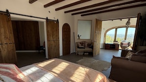 A spacious bedroom with a large bed, wooden beams, a view through a window, and a cozy sitting area with a sofa and a chair. - Villa 'Caroix' with Sea View, Wi-Fi and Air Conditioning (Xàbia)