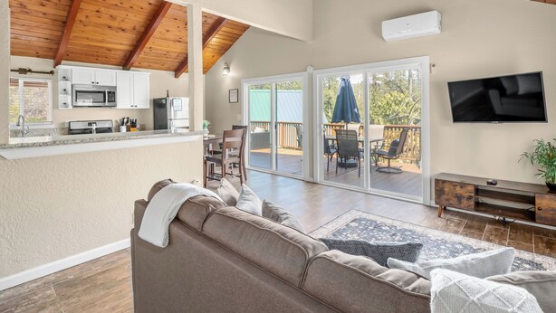A spacious living area with a kitchen, a large sofa, a TV, and a sliding glass door leading to an outdoor deck. - New! Arcade Games, Hot Tub,  Fire Pit, BBQ, 3:2 (Oakhurst)