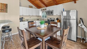 A kitchen with wooden ceiling beams, white cabinets, stainless steel appliances, and a dining table with chairs. - New! Arcade Games, Hot Tub,  Fire Pit, BBQ, 3:2 (Oakhurst)