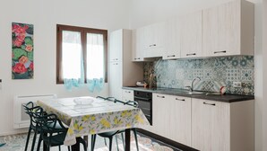A kitchen with light wooden cabinets, a tiled backsplash, a dining table with chairs, and a window with curtains. - Holiday Home 'La Vanedda' with Private Terrace, Wi-Fi and Air Conditioning (Partanna)