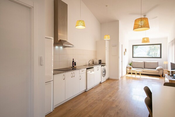 A modern kitchen with white cabinetry, a built-in oven, and a washing machine. There is a dining area with a sofa and a table.
