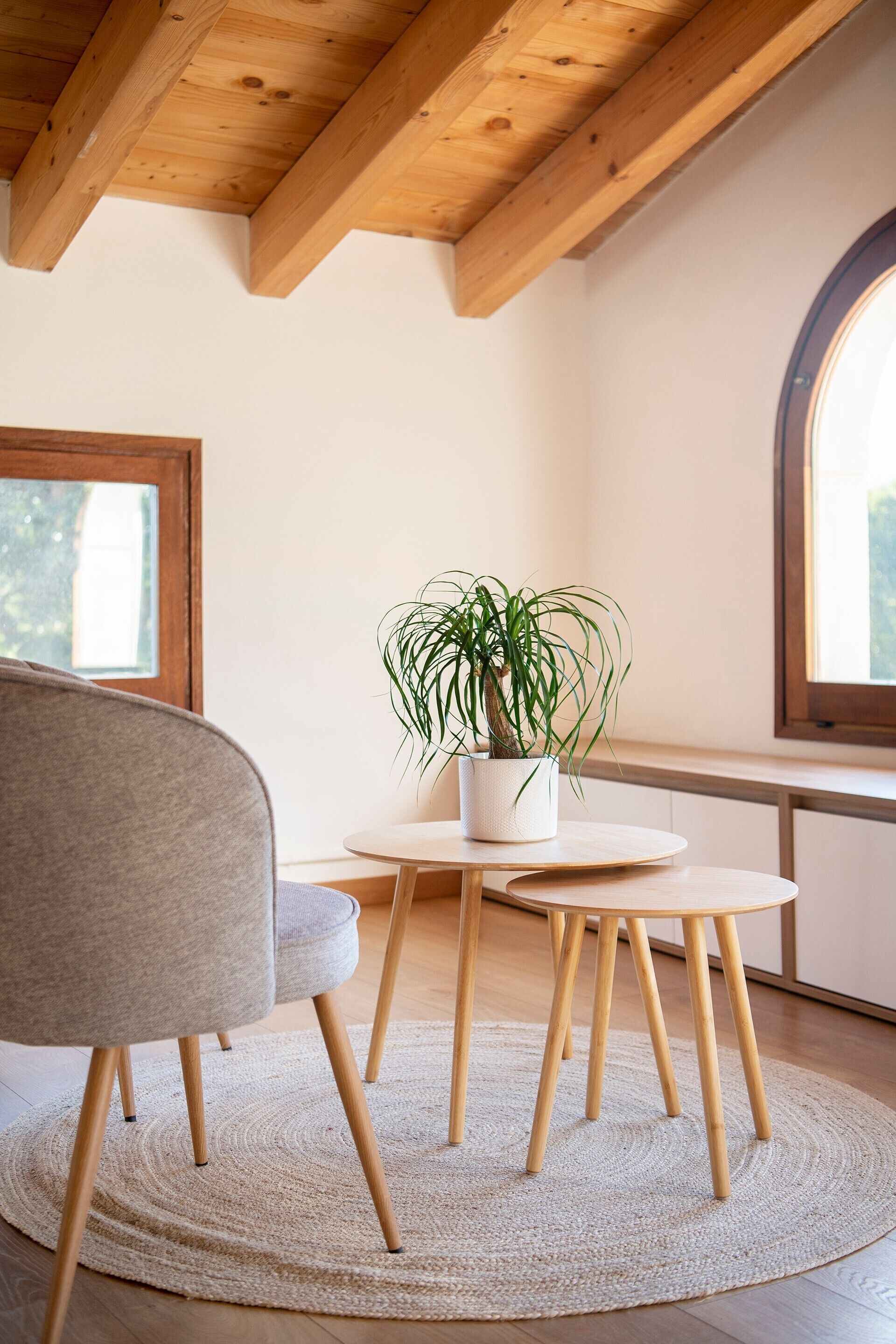 Una habitación con techo de madera, una mesa redonda con una planta en maceta, una silla gris y un ventanal grande.