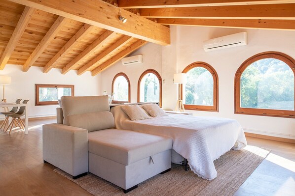 A bedroom with a large bed, a sofa, and a desk. There are arched windows and wooden beams on the ceiling.