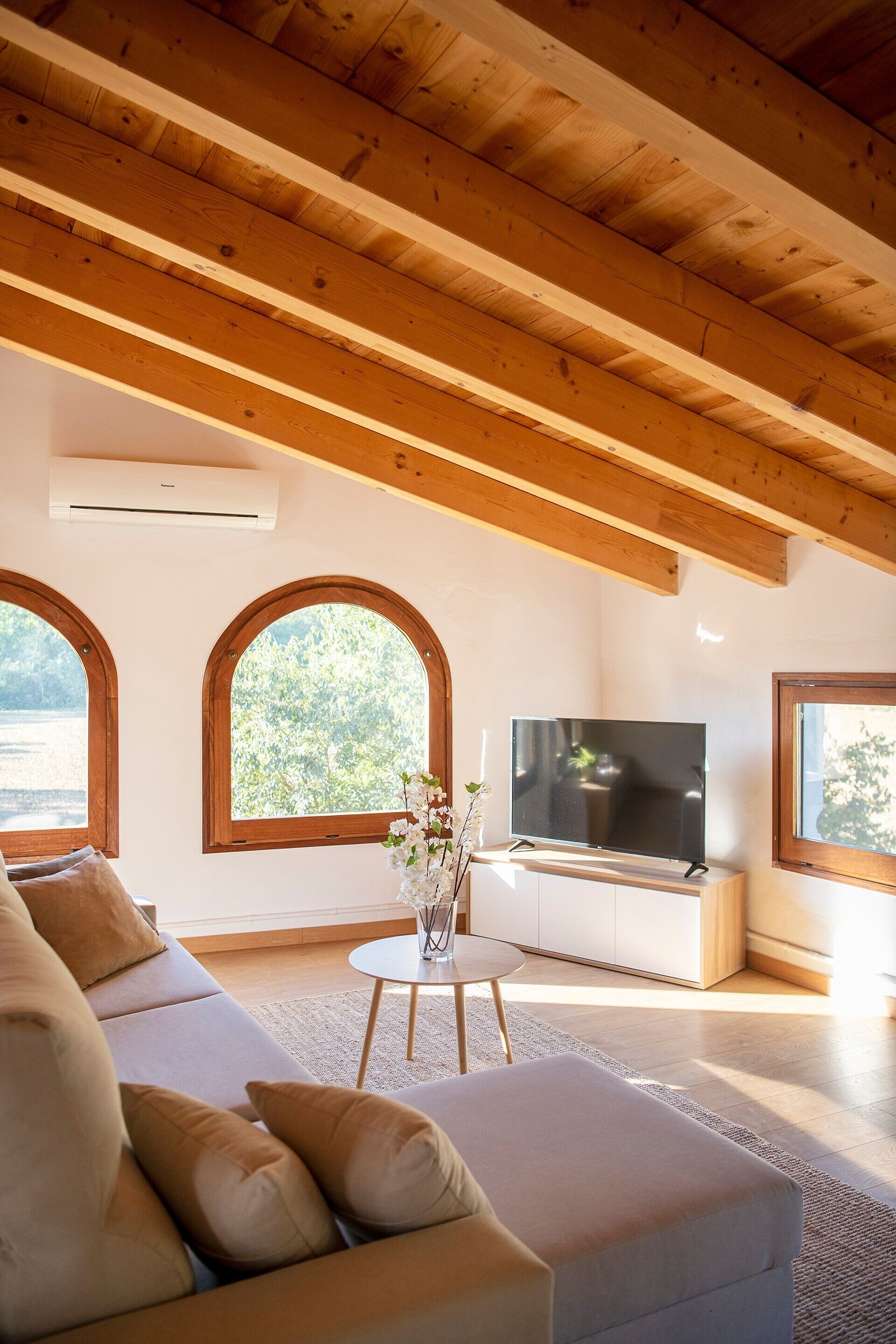 A modern living room with a wooden ceiling, a sofa, a TV, and large arched windows.