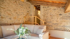 Une pièce aux murs de pierre, avec un escalier en bois, un canapé beige et une petite table sur laquelle est posée une plante.