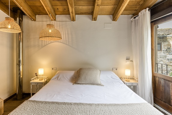 A bedroom with a large bed, two bedside lamps, a wooden ceiling, and a window with curtains.