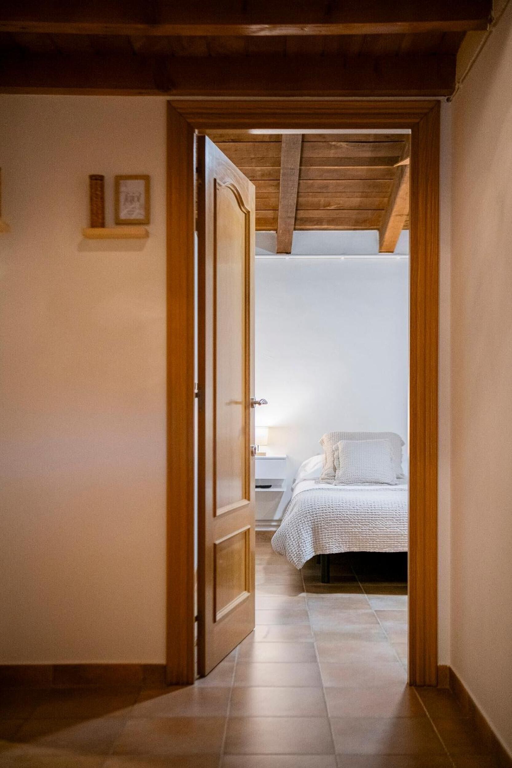 A narrow hallway leading to a bedroom with a bed, a nightstand, and a lamp. The room has wooden beams on the ceiling and a tiled floor.