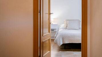 A narrow hallway leading to a bedroom with a bed, a nightstand, and a lamp. The room has wooden beams on the ceiling and a tiled floor.