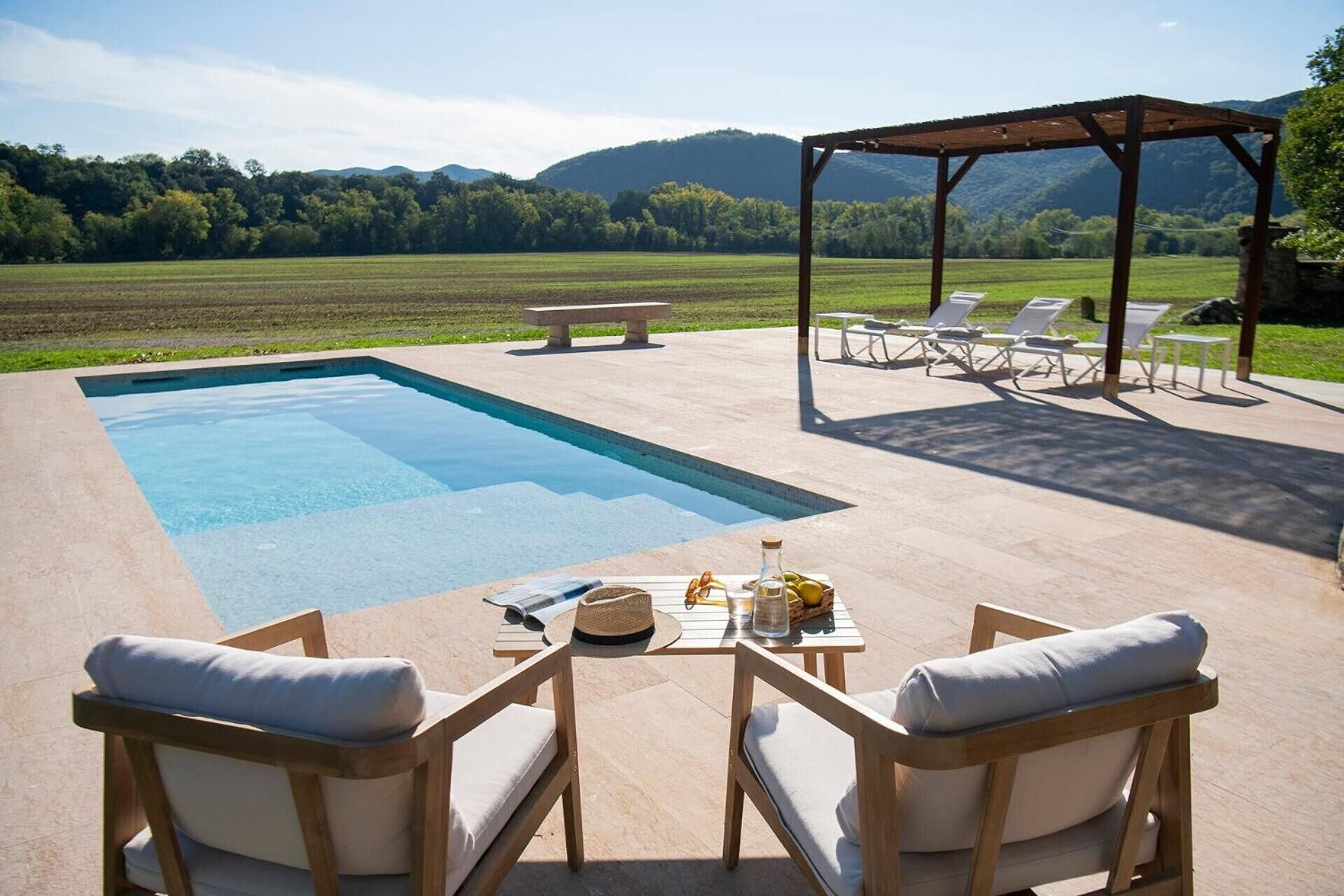 Una piscina con sillones de descanso y una mesa, rodeada por un patio con una pérgola y vistas al campo.