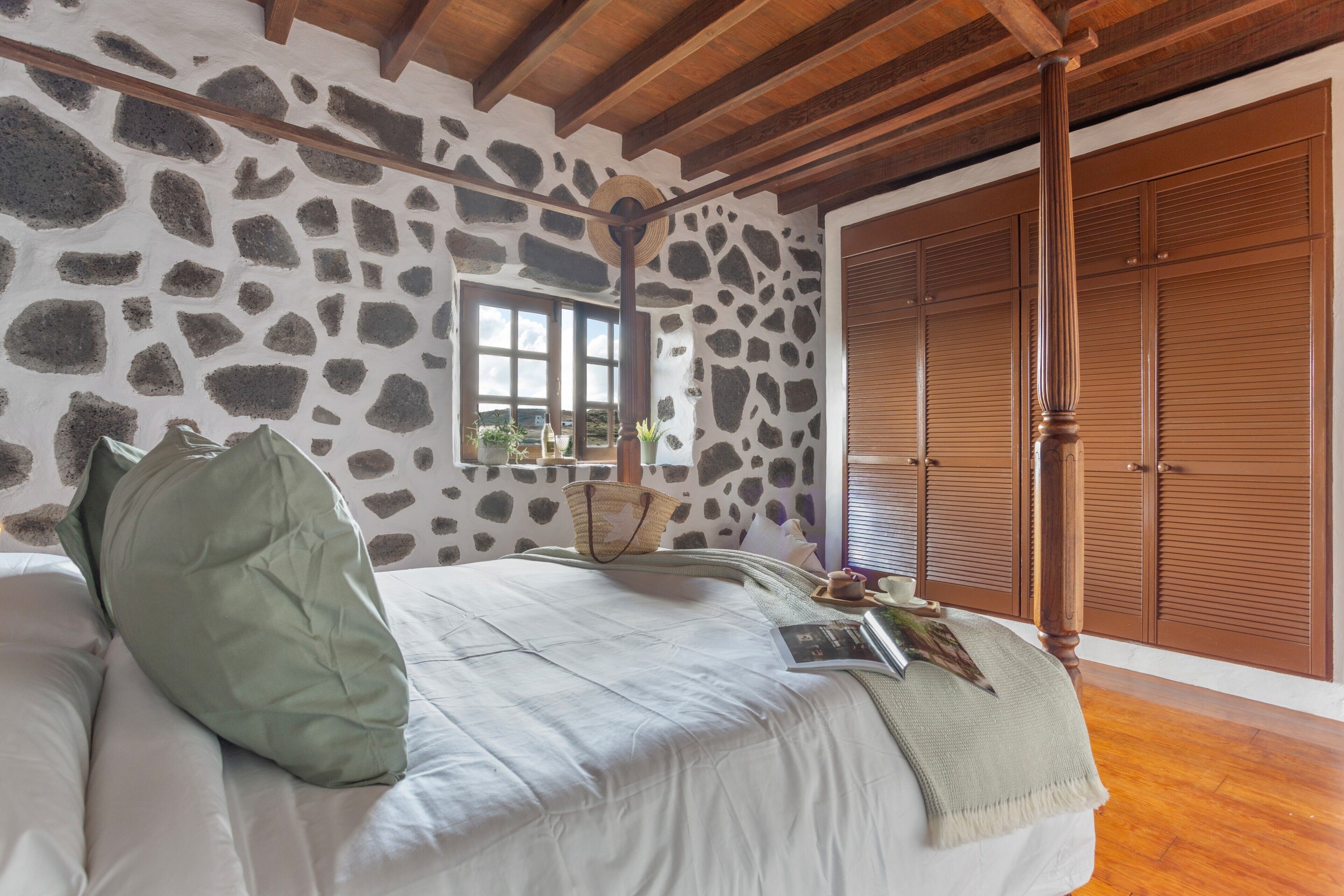 Une chambre avec un mur en pierre, des poutres en bois au plafond, un lit avec des oreillers, une fenêtre et une armoire.