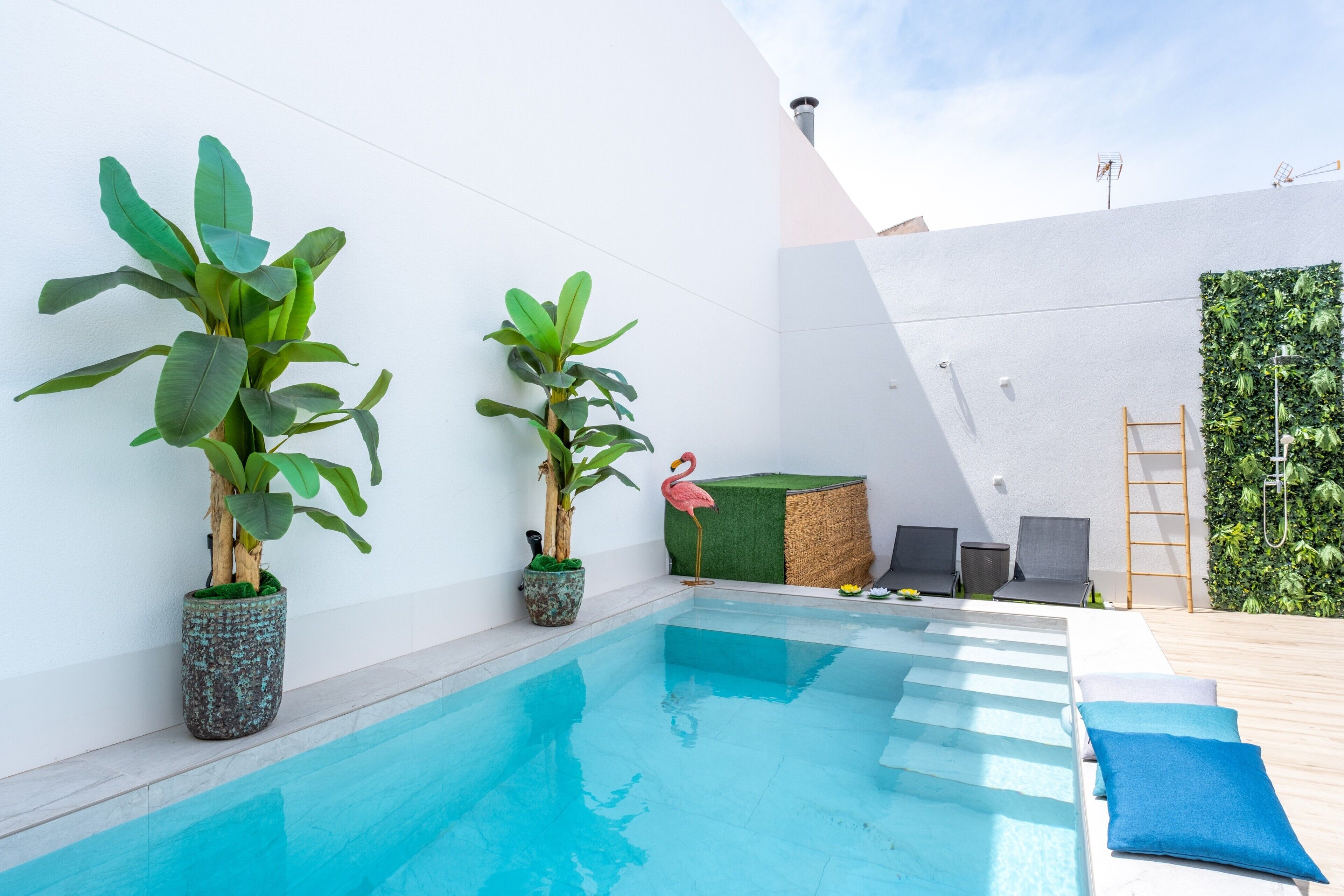 Un espace piscine extérieur moderne, avec une piscine à l’eau d’un bleu limpide, des plantes en pot et un petit coin salon agrémenté de coussins.