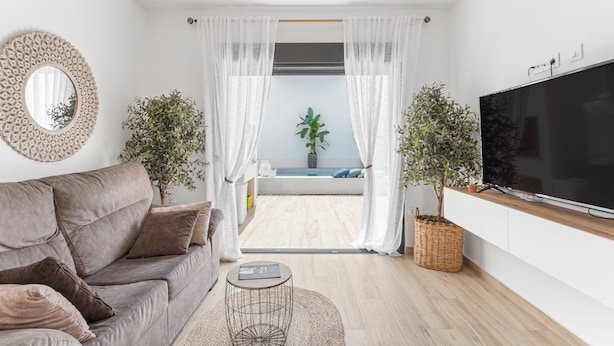 Un soggiorno moderno con un divano, una televisione, un tavolino e vista su un balcone con una pianta e una finestra.