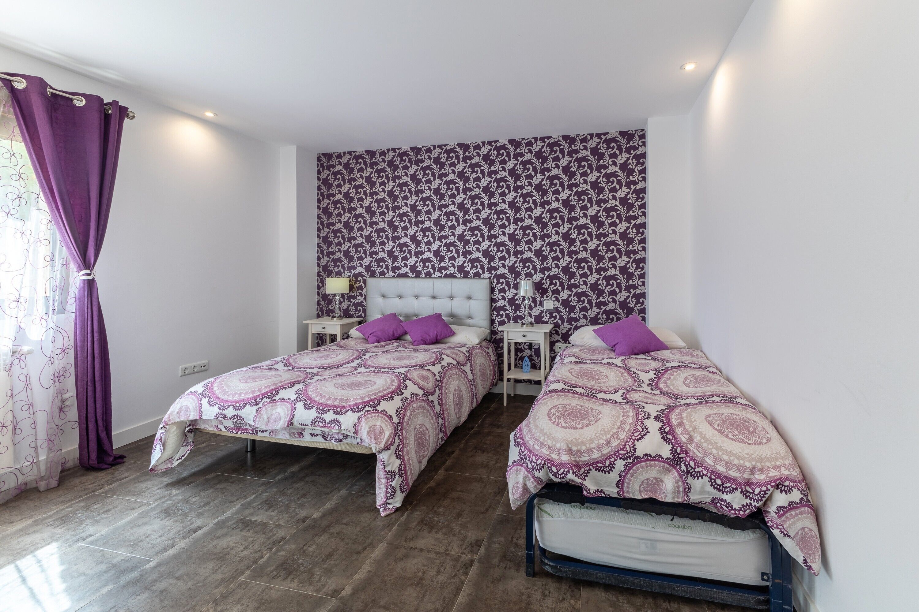 A bedroom with two beds, a patterned wallpaper, and a window with purple curtains.