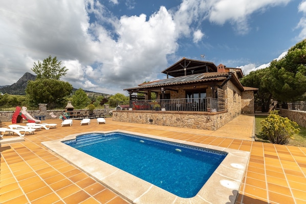 A pool area with a stone building, lounge chairs, and a mountain backdrop.