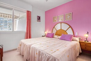 A bedroom with a bed, a nightstand, a lamp, a window with curtains, and a pink wall with framed pictures.