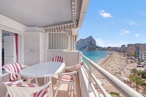 A balcony with a table and chairs, overlooking a beach and a mountain.