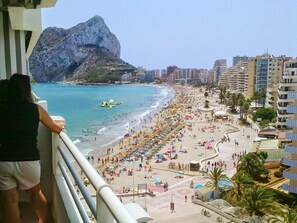 A beach with a rocky mountain in the background, a crowded beach area, and high-rise buildings along the shore.