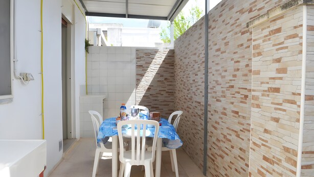 Un área de comedor al aire libre cubierta, con pared de ladrillo, sillas blancas y una mesa vestida con mantel azul.