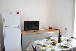 A dining area with a table set for two, a television on a stand, a refrigerator, and a bowl of fruit on the counter. - Holiday Home 'Casa A T Dell Orso X 6 Guests' with Private Garden (Torre dell'Orso)