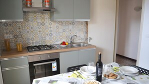 Una cocina con estufa a gas, pileta y alacenas. Se observa una mesa de comedor con platos, vasos y una botella.