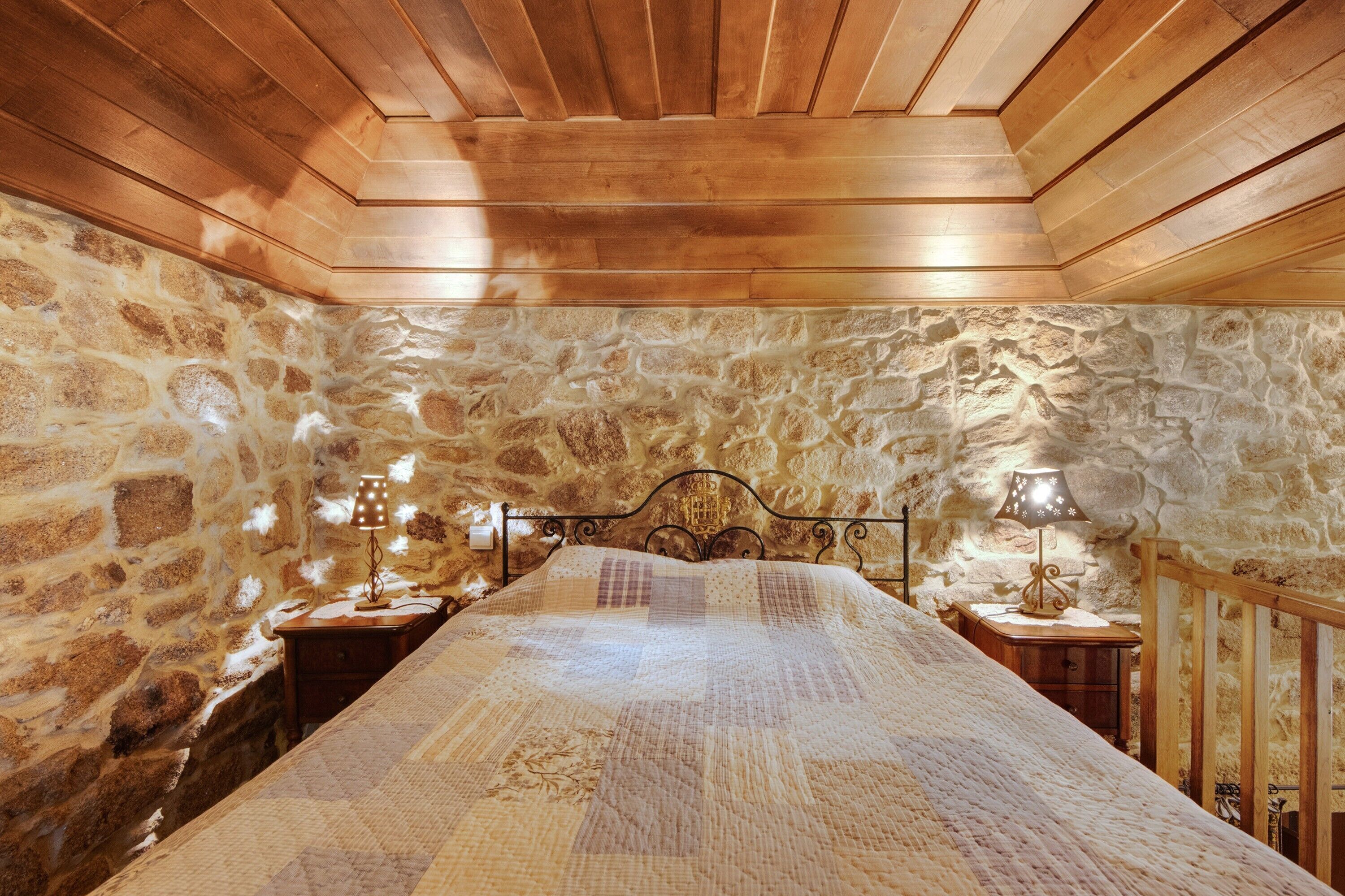A bedroom with a stone wall, wooden ceiling, a bed with a quilt, two bedside tables with lamps, and a wooden railing.