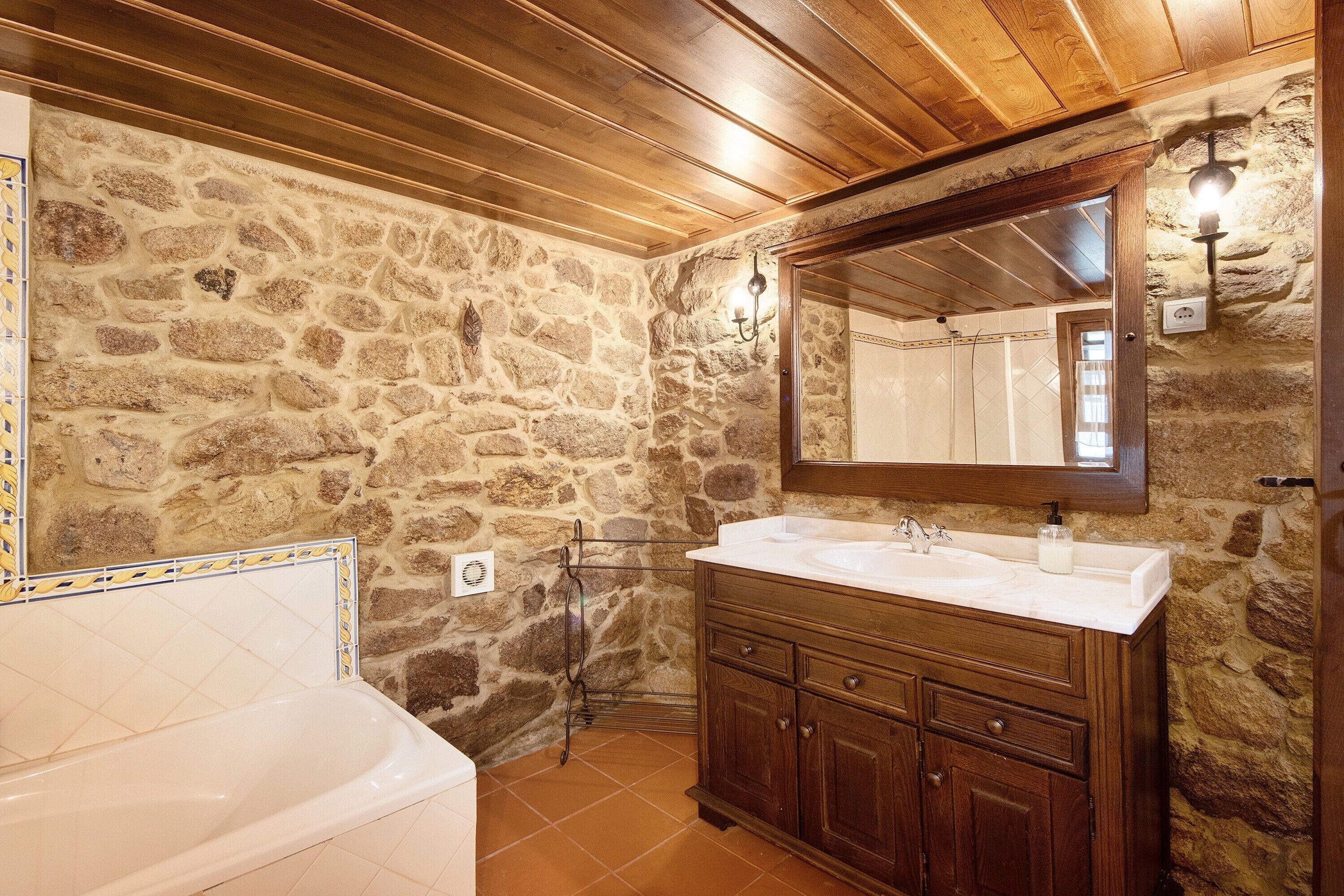 Un baño con pared de piedra, techo de madera, una bañera blanca y un tocador de madera con espejo.